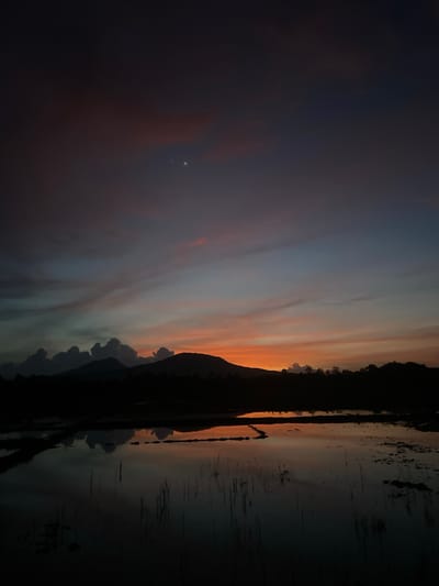 This image depicts a serene sunset scene over a rural landscape, likely in the vicinity of Khang Tanjong, Indonesia. The sky is a gradient of deep indigo at the top, transitioning to hues of soft pink, orange, and pale blue closer to the horizon, with thin wisps of clouds catching the last rays of sunlight. Two bright celestial bodies, possibly planets or very bright stars, are visible in the upper part of the sky. A silhouette of mountains forms a jagged line against the colorful sky, with darker, more irregular cloud formations nestled amongst them. In the foreground, a dark, reflective body of water, possibly a flooded paddy field or pond, mirrors the dramatic colors of the sky and the silhouetted landscape. Thin reeds or plant stalks emerge from the water in places. The overall atmosphere is tranquil and atmospheric, capturing the end of the day in a natural setting. There are no people or animals visible in the frame.
