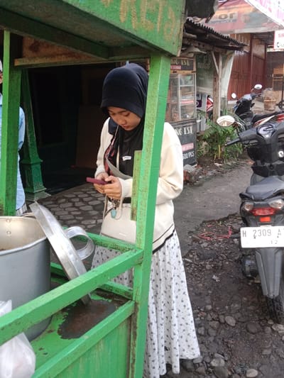 A woman wearing a black hijab, a white cardigan, and a white floral skirt is standing next to a green food cart. She is looking at a red mobile phone in her hands. The cart contains a large silver pot with a lid propped open. In the background, a motorcycle is parked on the street. A sign with the text "NG ISI DONG ISI" and "500/PCS" is also visible in the background. The motorcycle's license plate reads "H 2309 AT 12".