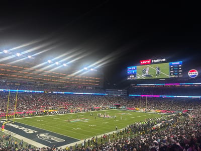 Levi's Stadium hosts a football game. Stadium lights shine brightly on the field, crowded with players and linesmen. Fans fill the stands on both sides. A large screen displays the game score (29-7) and a live football play. The Seattle Seahawks logo is painted in one end zone. A Pepsi advertisement is also visible.