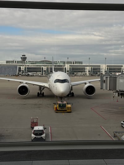 This image depicts an airport apron scene at Munich Airport (MUC) in Oberding, Germany. A large, white, twin-engine passenger aircraft, identified as a Lufthansa Airbus A350, is parked center-frame, nose pointed towards the viewer. Its left wing extends towards the foreground, and a jet bridge labeled "250/1" is connected to its right side.
A yellow pushback tug is positioned directly beneath the aircraft's nose gear. In the immediate foreground on the tarmac, a small red utility cart with a vertical pole and cone is visible to the left, and a white compact utility vehicle with small red flags mounted on its rear is parked behind it. Beyond the aircraft, multi-story modern airport terminal buildings stretch across the horizon. A distinctive tall, cylindrical air traffic control tower with a glass cab at its apex is prominent to the left of the central terminal structure. Several other smaller aircraft are visible on distant aprons. The sky is largely overcast with patches of lighter clouds. The scene is viewed through a large window, indicated by a dark frame at the top and a dark metal grating at the bottom of the image.
