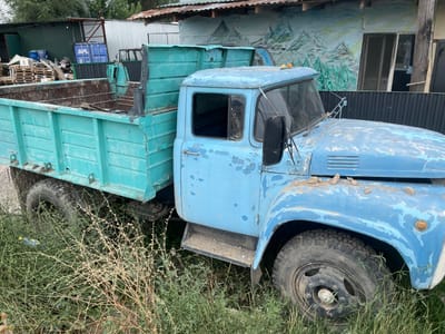 An aged, light blue flatbed truck is parked in an overgrown grassy area, suggesting it is disused. The truck's paint is peeling significantly, revealing rust underneath. The bed of the truck is a teal color and appears empty. Behind the truck, a corrugated metal fence runs horizontally, and beyond that, a building with a mural depicting a mountainous landscape is visible. Various objects, including wooden pallets, barrels, and a blue shipping container with the "APL" logo, are scattered around the area, indicating a storage or industrial yard. The environment is outdoors, with vegetation encroaching on the truck and its surroundings. No people are present in the image.