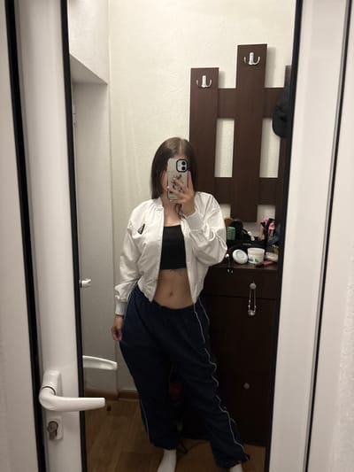An individual takes a mirror selfie indoors, obscuring most of her face with a smartphone held in her right hand. She has long, dark hair and light purple nail polish. She is wearing a black crop top, a white unzipped bomber jacket, dark blue track pants with a white stripe, and white socks. A navel piercing and a small tattoo on her left ribcage are visible. The reflection shows a domestic setting with white walls and a wooden floor. To the left of the individual is a white door frame with a white lever handle and a key inserted into the lock. To her right is a dark wooden coat rack with two silver hooks (one double, one single), on which a dark cap hangs. Below the coat rack is a dark wooden dresser holding various personal items, including hairbrushes, a white jar, a green-labeled jar, and a white electronic device. There is no visual information within the image to identify the location as Polena, Bulgaria.