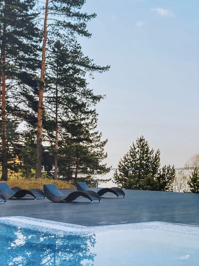 This image depicts an outdoor scene with a body of water in the foreground, reflecting the sky and surrounding trees. Behind the water is a dark, flat surface, likely a deck or patio, furnished with several dark, curved lounge chairs. Beyond the deck, a line of tall evergreen trees, primarily pines, stands against a pale blue sky with scattered clouds. In the background, a body of water is visible, with a distant tree line on the horizon. The location is identified as Novinki, Istrinsky District, Russia. There are no people visible in the image.