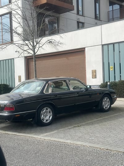A black Jaguar XJ sedan is parked on a light grey block-paved surface in Chatham, United Kingdom. The vehicle is oriented facing right, revealing its left side. A person, appearing to wear light-colored attire, is seated in the driver's seat.
Behind the vehicle, a modern multi-story building with a white facade and numerous dark-framed windows is visible. The ground floor features a large, horizontally ribbed, copper-colored roller shutter door. To the right of this door, a brass plaque bearing the text "2.8m" is affixed to the wall, next to a panel composed of light blue vertical slats displaying a yellow "H" symbol. A bare deciduous tree stands between the car and the building. Low green bushes are visible to the right of the vehicle, fronting the building. The immediate foreground consists of a darker grey asphalt surface with a concrete curb. The scene is illuminated by daylight, casting soft shadows.