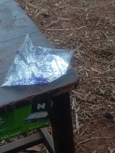 A clear plastic sachet containing water, labeled with "NATURAL" and "0.50L," rests on a dark, worn wooden table surface. Below the table, a green advertisement panel is partially visible, displaying a white Nigerian Naira symbol (₦), text related to "PRIZES" and "555 now," and a graphic depicting a dark, rectangular object. The scene is set outdoors in Egede, Nigeria, on dry, reddish-brown soil sparsely covered with scattered dry leaves, twigs, and plant debris. A small green sapling emerges from the soil in the background. No individuals are present.
