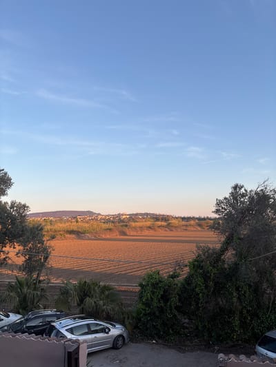 The image presents a landscape view taken from an elevated position, possibly a balcony. The right side of the image features a partially visible silver car parked near vegetation, including palm trees. A paved area suggests a road or driveway. To the left, the scene opens up to a view of agricultural land with distinct rows, indicating tilled soil. A line of buildings is visible in the distance, suggesting a populated area. The sky above transitions from a pale blue to a light orange hue, indicative of either sunrise or sunset. The presence of trees along the edge of the frame further frames the scene. The setting appears to be Giugliano in Campania, Italy, based on the landscape and architectural characteristics.