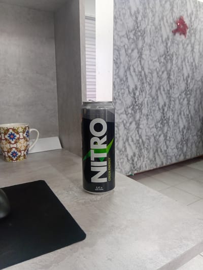 The image depicts a Nitro Green Energy drink can placed on a light grey countertop. To the left of the can is a colorful mug with a patterned design. In the background, there is a textured wall and a white partition. The overall scene appears to be an indoor setting, possibly a desk area. There is no indication of Tokmok City, Kyrgyzstan, or any specific location.