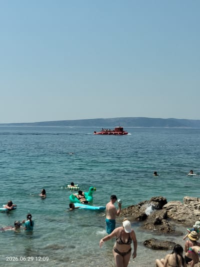 This image depicts a sunny day at a beach in Krk, Croatia. The water is clear and blue, with several people enjoying various activities. In the background, a red submarine-like boat is visible on the sea, with people on its deck. Closer to the shore, individuals are swimming, floating on inflatable toys, and relaxing on the pebbly beach. One person is holding a white bucket, appearing to scoop water from the sea. Rocks are present along the shoreline, with some waves crashing against them. The sky is clear and light blue. The timestamp in the lower left corner indicates the image was taken on June 29, 2026, at 12:09.