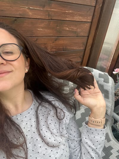 The image shows a woman lying down, possibly on a deck or patio, with a wooden plank surface to her left. She's wearing a light grey sweater with small black polka dots and a beige smartwatch. She has long, dark brown hair, and is wearing glasses with black frames. Her hand is raised, holding a strand of her hair. A grey and white patterned cushion is visible behind her hand. In the background, there is a potted plant with pink flowers. The location is impossible to determine from the image.