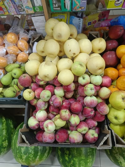 This is a waist-level, eye-level, medium shot of a fruit stall in a market in Gulbakhor, Uzbekistan. In the foreground, several watermelons are visible, with their striped green rinds taking up the bottom left and bottom right of the frame. Above the watermelons, and to the right, are several crates filled with various fruits. The largest crate, in the center of the image, is overflowing with a mix of red and yellow apples, as well as some pears. To the right of this crate, a smaller crate is visible, also filled with red and yellow apples. Further to the right, oranges are piled high. The background shows shelves stocked with packaged goods, including boxes of tissues, snacks, and other grocery items. The lighting is bright and even, suggesting an indoor or shaded outdoor market setting.