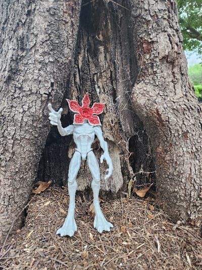 A Demogorgon action figure, with a red petal-shaped head, rests in the crevice between two halves of a fallen tree trunk. The figure is white with elongated limbs and clawed hands and feet. The surrounding environment includes the rough texture of the tree bark, dry leaves, twigs, and a faint spiderweb. The scene is outdoors, possibly in a wooded area.