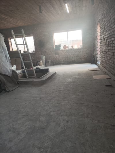 The image depicts the interior of a partially constructed or renovated building, likely a garage or workshop, located in Tselina, Russia. The space has a concrete floor and brick walls. A wooden ceiling is visible, with exposed beams and a fluorescent light fixture. Natural light streams in through two large windows on the far wall, which is made of brick.
In the foreground, a square concrete pit with a raised edge is present. A silver ladder leans into this pit. Various objects are scattered around, including what appear to be car batteries, a toolbox, and possibly car parts covered in plastic sheeting on the left side of the frame. To the right, near the wall, are some stacks of materials, including what look like buckets and perhaps some concrete blocks. Two wooden planks are lying on the floor near the right wall. The overall impression is one of a work in progress, with an unfinished feel to the space. There are no people visible in the image.