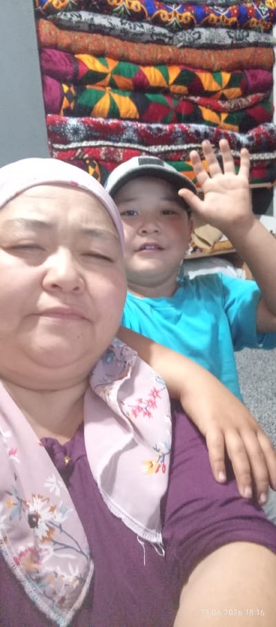 A medium shot captures a woman and a child posing for a selfie. The woman, in the foreground and to the left, is wearing a pink headscarf and a purple shirt with a floral patterned scarf. She has her eyes closed and appears to be smiling. Behind her and to the right is a child wearing a blue t-shirt and a grey baseball cap. The child is smiling and waving at the camera with their right hand. The background features a wall covered with a stack of colorful, patterned textiles, likely blankets or cushions. A date stamp at the bottom right reads "23.06.2026 18:16." The overall impression is one of a casual, indoor family portrait taken in Chon-Kara, Kyrgyzstan.