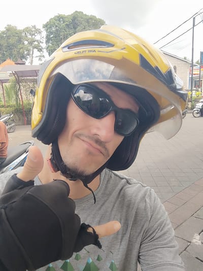 A fair-skinned male is taking a selfie. He is wearing a yellow motorcycle helmet with a clear visor, and dark sunglasses. He has a mustache and a light beard. He is wearing a gray t-shirt and black fingerless gloves. He is giving a thumbs-up sign with one hand and pointing with the other. The background shows a street scene in Bedulu, Indonesia, with trees, buildings, and parked motorcycles. The ground is paved with rectangular stones. Overhead wires are visible.