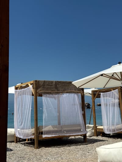 The image depicts a serene beach scene on a clear, sunny day in Vrba, Montenegro. The sky is a vast, uninterrupted expanse of bright blue, with a faint, thin white contrail from an aircraft visible in the upper central portion. The horizon line is sharply defined where the sky meets the calm, turquoise-blue water of the sea.
The foreground consists of a light-colored pebble beach. Two wooden-framed cabanas, or daybeds, are prominently featured on the beach. Each cabana has a rustic wooden structure, topped with a textured, woven, light brown roof material. White, sheer fabric curtains hang from the sides of the cabanas, gently billowing in the breeze, revealing light-colored mattresses or cushions within. Scattered on the beach and near the cabanas are white, soft beanbag-style cushions.
On the right side of the frame, a large white patio umbrella with a wooden frame and a dark finial is partially open, casting a distinct shadow. Another smaller white umbrella is partially visible behind the leftmost cabana.
The sea exhibits a clear turquoise hue. White buoys are visible floating on the water, marking a designated area. On the far left, a person's head is visible, partially submerged in the water. In the mid-ground, towards the right, large dark rocks protrude from the sea, and a small watercraft with an engine and a dark figure aboard is discernible near these rocks. The overall impression is one of leisure and relaxation under bright natural light, with strong shadows indicating the sun's high position. A thick, dark wooden post frames the left edge of the composition.