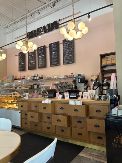 The image shows the interior of a cafe with a wooden counter, shelves stocked with bags and cups, and menu boards hanging on a pale pink wall. There are pastries displayed in a glass case on the left. A large chandelier with multiple globes hangs from the ceiling. In the foreground, there are chairs and a table. The cafe is well-lit and appears to be open for business. The text on the menu boards suggests it is a location in the city of Vaughan, Canada, as the name "Bready" is prominent and the menu items are typical of a cafe or bakery.