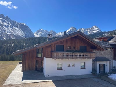 The image depicts a chalet-style house in a mountainous region, likely Sappada, Italy. The house has a distinctive architectural style with a wooden upper story and a white stucco lower story. There is a balcony with wooden railings, and some greenery is visible on the balcony. Windows are visible on both levels, some with closed shutters. The house has a sharply angled roof, and a chimney is present.
In the background, there are snow-capped mountain peaks and a dense forest of evergreen trees. The sky is clear blue with a few scattered clouds, suggesting a sunny day. The foreground shows a paved area leading up to the house. To the right, part of another building with a stone exterior is visible, along with some melting snow patches. The overall scene is characteristic of a mountain resort town.