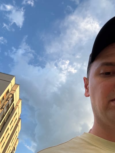 The image is a low-angle, upward-facing shot, likely a selfie. It features the upper part of a person's face, including an eye and ear, framed by a dark baseball cap. The person is wearing a light-colored t-shirt. The background consists of a sky with a mix of blue and gray clouds, suggesting a day with variable weather. A section of a modern building with light-colored facade and numerous windows is visible in the lower right portion of the frame, partially obscured by the person's t-shirt. The overall impression is casual, possibly taken outdoors on a cloudy but bright day. There is no visible text in the image. The location is identified as Koltsovo, Russia.