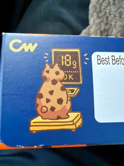 The image shows a close-up of a blue box with a cartoon illustration. On the left side of the box, there is a yellow logo that appears to say "CW". To the right of the logo, there is a cartoon of a dalmatian-patterned cat sitting on a yellow scale. Above the cat, there is a computer monitor displaying "18g" and "OK". To the right of the illustration, there is a white label with the words "Best Befo" printed on it, suggesting it is part of a "Best Before" date. The box appears to be a food product. The image does not provide any information about the location in Tofta, Sweden, nor does it depict any people or specific actions.
