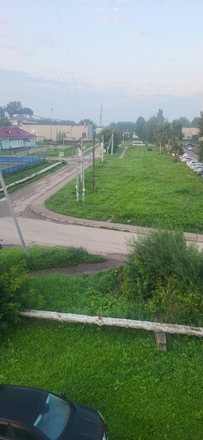 The image depicts a view from above, looking down on a street and a grassy area in the city of Gamovo, Russia. The street is paved and curves to the left. Several pedestrian crossings are marked on the street with yellow and white signs. Utility poles with wires are visible along the street. A white van is parked on the right side of the road, and a line of parked cars extends further down the street. The grassy area beside the street is lush and green, with scattered trees. In the foreground, a section of an insulated pipe runs horizontally across the image, supported by concrete blocks. A portion of a black car is visible at the bottom left of the frame. The sky above is pale blue with some wispy clouds. Buildings, including a green and white structure and a larger beige building, are visible in the background to the left. A red antenna or tower is also present in the distance. The scene appears to be an everyday urban environment.