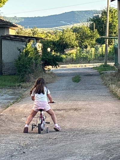 A young girl with long brown hair is riding a small pink bicycle with training wheels down a dirt road in Enevo, Bulgaria. She is wearing a white t-shirt and pink shorts. The road is lined with trees and houses. In the background, there are hills. The sun is setting, casting a warm glow on the scene.