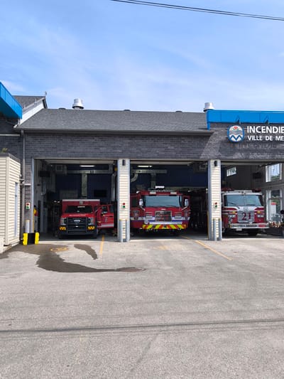 The original image depicts the exterior of a fire station located in Mercier, Canada, under a clear blue sky with scattered clouds. The building features a dark gray brick facade with large, open garage bays. Three red fire trucks are visible, two parked in the garage bays and one partially obscured. The fire truck on the far right has the number "21" prominently displayed on its front. A blue awning extends over the top of the garage bays. A circular emblem with the words "INCENDIE" and "VILLE DE MERCIER" is affixed to the upper right portion of the building, near the blue awning. The foreground shows a paved area with yellow painted lines and a dark, amorphous stain on the ground, possibly from a fluid leak. Yellow traffic cones and a red and green signal light are also present near one of the garage bays. The overall scene is orderly and suggests a functional emergency services facility.