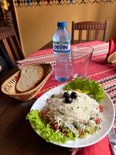 The image displays a close-up, overhead view of a table set for a meal, likely in a restaurant. The central focus is a large white plate filled with a fresh salad, resembling a Shopska salad. It features a base of vibrant green lettuce, diced tomatoes, and cucumbers, all generously covered with a mound of grated white cheese. Three black olives are positioned on a small skewer alongside a tiny yellow pickled pepper, resting on the cheese, which is also sprinkled with green herbs.
Adjacent to the salad, a transparent plastic bottle of "DEVIN" branded water with a blue cap is visible. The label clearly shows "DEVIN" and the Cyrillic text "ИЗВОРНА ВОДА" (meaning "Spring Water"). An empty clear drinking glass stands nearby. A small, woven wicker basket contains several slices of bread, including both lighter and darker varieties.
The table is covered with a decorative red tablecloth adorned with colorful, intricate geometric patterns, reminiscent of traditional folk textiles. In the background, a dark wooden chair is partially visible, and the wall behind it is a plain, light brown color with a complementary patterned border near the floor. The scene suggests a prepared meal awaiting consumption in an inviting, rustic indoor setting. No individuals are present in the frame.