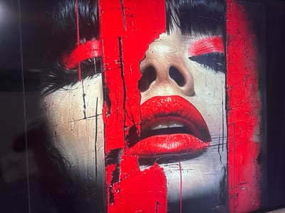 The image displays a close-up of a person's face, with heavy red and black paint applied in a graphic, abstract style. The subject's mouth is prominently featured, with bright red lipstick and a tongue slightly visible. The eyes are partially obscured by red paint, with visible eyelashes. The background is dark and appears to be a digital screen or monitor. There is no discernible urban environment or location within Krāslava, Latvia. The image focuses solely on the artistic representation of the face.