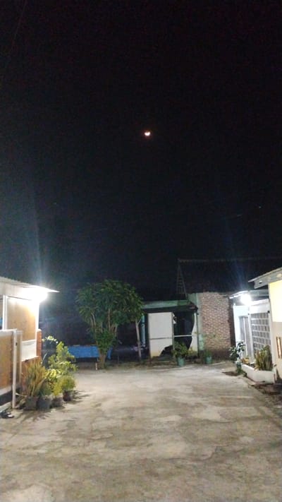 The image depicts a nighttime scene on a paved pathway in Banjarsari, Indonesia. The sky is dark, with a crescent moon visible in the upper center. To the left, a building with a bright light emanating from its side is partially visible. In the middle ground, a tree with dense foliage stands, and behind it are structures with brick walls and tiled roofs, illuminated by artificial lights. Potted plants are placed along the pathway, near the buildings. The pathway appears to be made of concrete, with some visible cracks and discoloration. The overall impression is a quiet residential or communal area at night.