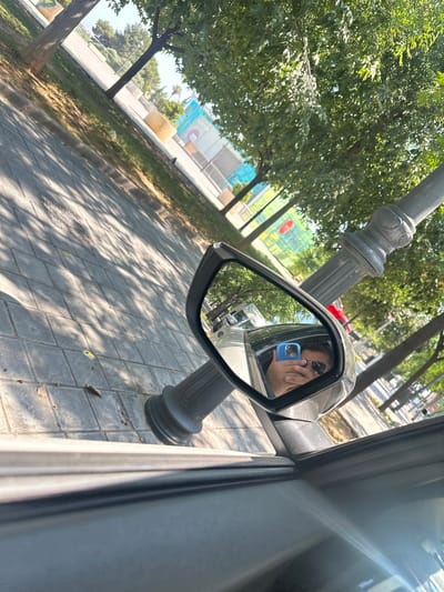 The image shows a reflection in a car's side mirror of a person holding a blue smartphone and wearing sunglasses. The person appears to be taking a selfie. The background of the reflection shows trees and buildings. The car is parked on a paved surface, possibly a street or walkway, with a lamppost visible in the foreground. The location is identified as Benimàmet-beniferri, Spain, suggesting an urban or suburban environment. The overall lighting indicates it is daytime, likely sunny, with shadows cast on the ground.