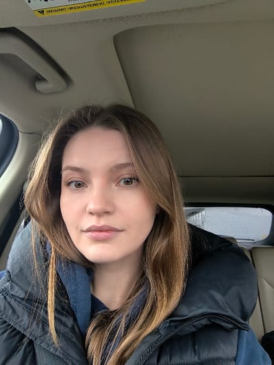 A female individual is depicted in a close-up, front-facing portrait, positioned within the passenger compartment of a vehicle. She has light brown hair, brown eyes, and a neutral facial expression, looking directly at the camera. She is wearing a dark blue hooded garment beneath a dark blue or black puffer vest. Above her head, a light-colored vehicle ceiling, an overhead grab handle, and a yellow warning label with inverted text are visible. Through the rear window of the vehicle, a blurred outdoor scene featuring indistinct structures and a rear windshield wiper arm is visible. The seat upholstery visible behind her is a light beige or cream color. The precise location within Illintsi, Ukraine, cannot be ascertained from the visual evidence presented in this image.