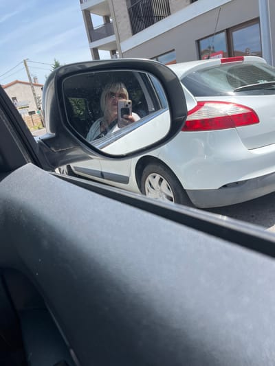 The image captures a scene from the interior of a vehicle, looking towards the passenger-side exterior on a sunny day. The foreground shows a dark, textured dashboard or door panel.

Dominating the midground is the vehicle's side-view mirror, reflecting the driver. The reflection shows an adult individual, likely female, with blonde hair and sunglasses, holding a light-colored smartphone with a dark case, actively taking a selfie. She is wearing a light-colored top.

To the right of the mirror, the rear section of a white Renault Mégane II hatchback is visible, parked or stopped parallel to the camera's vehicle. The car displays a red and white taillight assembly, a rear window wiper, and a roof-mounted antenna. The lower portion of its rear bumper is dark grey, with a visible indentation or minor damage on the right side. Faint reflections of a building with red signage are visible in the Mégane's rear window, including what appears to be part of the word "OUVERT" (French for "OPEN").

The background, visible outside the mirror and the white car, depicts a street scene in Saint-Just-Saint-Rambert, France. A multi-story building with brown balconies stands prominently. To its left, a light-colored, older-style house with a tiled roof is partially visible. A utility pole with multiple wires and a street signpost are present. The street signpost clearly displays white rectangular signs indicating directions, including "SAINT-RAMBERT CENTRE VILLE", "ST ETIENNE", and "ANDRÉZIEUX BOUTHÉON", definitively confirming the location. A circular regulatory road sign (red border, white center) is also attached to the pole. The sky is clear and blue with minimal cloud cover.