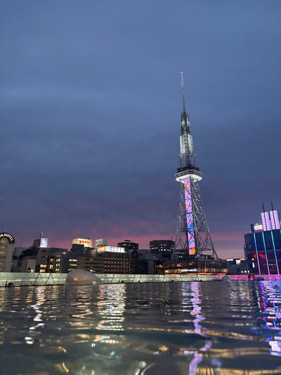 This image captures a twilight scene in Nagoya, Japan, likely from a rooftop vantage point. The most prominent feature is the Nagoya TV Tower, its lattice structure illuminated with vibrant, shifting colors. The sky is a deep indigo transitioning to a soft pink and orange horizon, indicative of a sunset or afterglow.

In the foreground, a body of water, possibly a swimming pool or reflective surface, mirrors the colorful lights of the city and the tower. The ripples on the water's surface distort the reflections, creating abstract patterns of light.

Behind the tower, a cityscape unfolds with various buildings. Some display illuminated signage, including "KOTSUGEN" and "DAIICHI KANKO." The architecture suggests a commercial or entertainment district. To the right of the tower, another skyscraper glows with purple and blue neon lights.

There are no visible people in the immediate foreground. However, in the far right background, near the glowing skyscraper, a few figures can be faintly discerned, suggesting the presence of people in the urban environment. The overall atmosphere is one of modern urban beauty, enhanced by the dramatic interplay of natural twilight and artificial illumination.