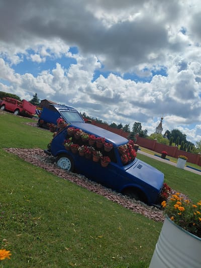 In Kovalivka, Ukraine, an outdoor scene features two vintage vehicles repurposed as planters. A blue car is positioned at a downward angle, with its interior and exterior adorned with numerous potted flowers. In the background, a red car, also filled with flowers, is visible on a slight incline. The cars are situated on a grassy area interspersed with red gravel. Beyond the cars, a brick wall encloses a garden area with trees and a distant church with a golden dome. The sky is filled with dramatic clouds.