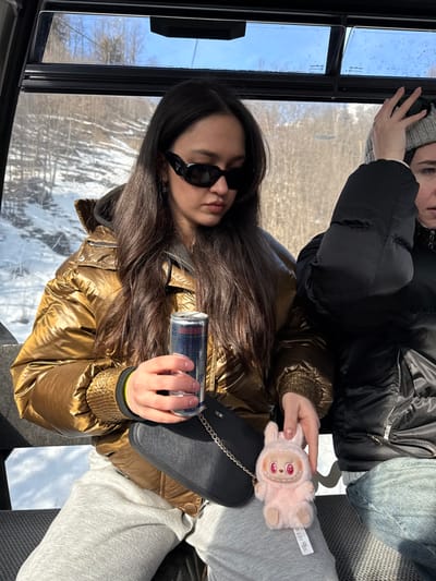 The image captures two individuals seated inside what appears to be a cable car or gondola, traveling through a snowy, mountainous landscape in Alagna Valsesia, Italy. The setting suggests a bright, sunny winter day.
In the foreground, a young woman with long dark hair is visible, wearing a shiny gold puffer jacket and black rectangular sunglasses. She holds a red and silver Red Bull can in her right hand. Her expression is neutral, and she appears to be looking towards the viewer. She is also wearing light grey sweatpants, and on her lap, next to a small black shoulder bag with a silver chain, rests a distinctive light pink furry keychain or toy with red eyes and a small mouth. A fabric tag, partially visible on the toy, contains some unreadable text.
Behind her, another individual is partially visible, wearing a grey knitted beanie and a black puffer jacket. Only the side of her face and one hand are seen, and she appears to be looking out the window or slightly away.
Through the large windows of the cable car, a clear blue sky and snow-covered trees on mountain slopes are visible, indicating a high-altitude winter environment. The sunlight illuminates the interior and reflects off the windows. The interior structure includes dark framing around the windows and patterned grey seating. The overall scene depicts a relaxed journey through a picturesque winter landscape.