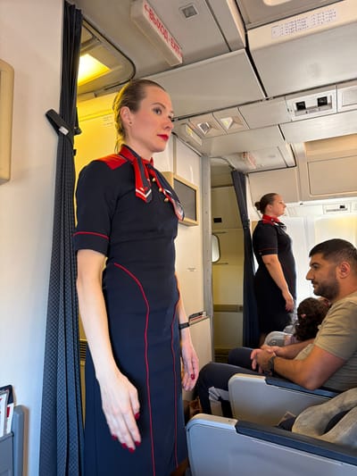 Inside an aircraft cabin at Dalaman, Turkey, two female flight attendants in dark blue dresses with red piping and scarves are positioned among seated passengers. In the foreground, a light-complexioned female flight attendant with her hair pulled back and wearing red lipstick stands upright, facing left. She has red nail polish and a black wristband on her left wrist. Behind her, a dark grey patterned curtain with a checkerboard texture is pulled open, revealing a brightly lit yellow compartment. An overhead sign displays "ВЫХОД - EXIT" in red and white lettering, along with a "No Stowage" sign below it. Further into the cabin on the right, a second female flight attendant, of a heavier build and similarly uniformed, stands facing right. In the right foreground, a male passenger with dark hair and a beard sits in an aisle seat, wearing an olive-green t-shirt and a black watch on his left wrist. His hands are clasped, and he is looking towards the main flight attendant. A child with dark, curly hair is partially visible seated next to him. Overhead panels feature passenger service units, including call buttons and a sticker with Braille text. The overall scene is illuminated by interior cabin lighting and the warm light from the open compartment.