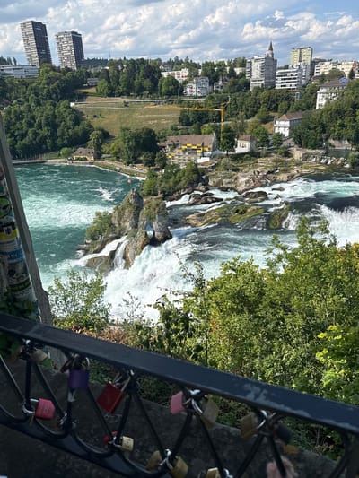 This media file captures a scenic view of the Rhine Falls in Nohl, Switzerland. The image is oriented vertically, looking down towards the powerful waterfall. The main subject is the majestic waterfall itself, with its turbulent white water cascading over large, moss-covered rocks. The water is a vibrant turquoise green where it pools at the base of the falls.
Surrounding the falls, lush green trees and vegetation line the banks. In the background, a city skyline is visible with several buildings, including apartment blocks and older structures. A yellow construction crane is also present, suggesting ongoing development or maintenance in the area. The sky is a bright blue with scattered white clouds, indicating pleasant weather.
In the foreground, a dark metal railing is visible, adorned with numerous colorful love locks of various sizes and hues, including pink, red, purple, and yellow. These locks, attached to the railing, are a common symbol of love and commitment. Some of the locks have numbers or initials inscribed on them.
The overall scene evokes a sense of natural beauty and grandeur, combined with the human touch of the love locks. The lighting suggests it is daytime, likely midday or afternoon. The image conveys a feeling of awe at the power of nature and the romantic sentiment associated with the love locks. Some sticker-like elements with illegible text are visible on a pole or structure near the bottom of the frame.