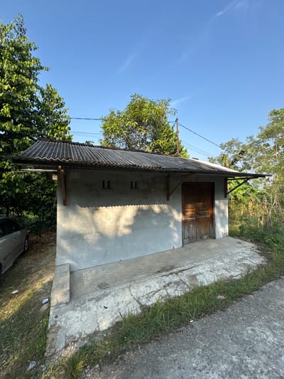 A one-story, weathered concrete building with a corrugated metal roof stands under a clear blue sky in a rural area, possibly Alian, Indonesia. The building has a single set of wooden double doors on its front facade, flanked by two small, square windows. A concrete path leads up to the doors. To the left of the building, a silver car is parked partially visible. Lush green trees and vegetation surround the structure, and power lines stretch across the sky above. The scene appears to be bathed in the warm light of late afternoon or early morning.