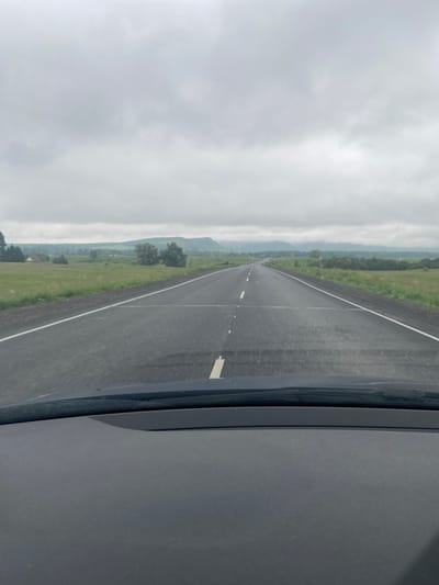 The image shows a view from inside a car looking out at a two-lane asphalt road stretching into the distance. The road is marked with white lines. The sky is overcast with gray clouds. On either side of the road are green fields with scattered trees and some distant hills. A portion of the car's dashboard and windshield are visible in the foreground. The scene appears to be a rural or semi-rural setting, and there is no indication that this scene is in the city of Arkhangelskoye, Russia.