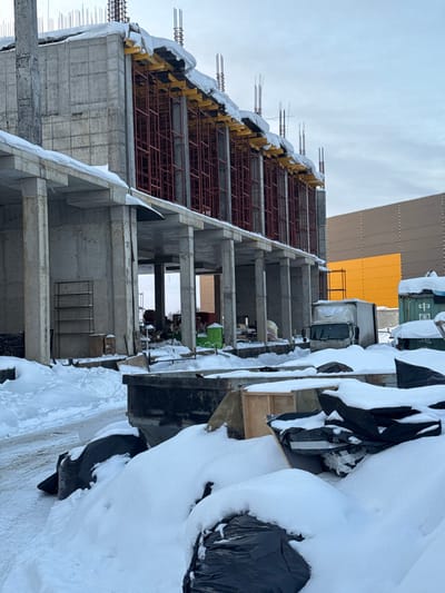 The image displays a winter construction site located in L'vovskiy, Russia, under an overcast, light grey sky. The scene is heavily covered in white snow.
In the foreground, large piles of snow conceal dark debris, some wrapped in black plastic sheeting. A dark-colored, open-top dumpster is partially visible in the center, filled with snow and various materials. To its right, wooden construction elements are partially obscured by snow.
The midground is dominated by an unfinished multi-story concrete building structure. Its exposed concrete framework includes numerous columns supporting multiple floor slabs. The upper visible floor features red metal scaffolding supporting yellow formwork panels along a significant portion of its length, with exposed rebar protruding from the top edge, indicating ongoing construction. Snow accumulates on the concrete slabs and formwork. Beneath the elevated section of the building, a green portable toilet, a white box truck, and a blue shipping container marked with Chinese characters are discernible amidst various construction materials and equipment. A metal shelving unit is visible under the structure on the left.
In the background, a large, rectangular building with a brown paneled facade featuring a prominent yellow section is visible to the right of the concrete structure. No individuals are present in the scene.