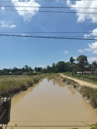 The image depicts a drainage canal in Keudepanteubreuh, Indonesia. The canal is filled with muddy brown water. It is flanked by vegetation and a dirt path. In the background, there are trees, rice paddies, and some buildings. Electrical wires stretch across the top of the image. The sky is blue with scattered clouds. In the foreground, a concrete structure is visible.