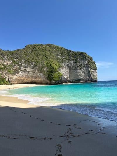 The image shows a scenic view of Kelingking Beach, located in Bunga Mekar, Nusa Penida, Indonesia. A large, verdant cliff dominates the left side of the image, its textured rock face partially covered in vegetation. Adjacent to the cliff is a narrow sandy beach, where a few people can be seen. The turquoise water meets the beach with gentle waves. Footprints are visible on the darker sand in the foreground. A clear blue sky completes the backdrop.