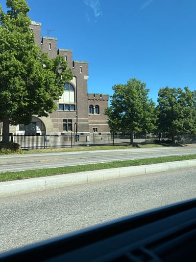 The image shows a view of the Helsingborg Fortress (Kärnan) and its surroundings in Helsingborg, Sweden. The fortress, made of brick, has a castellated appearance with stepped battlements and arched windows. Several large, green trees are situated in front of and to the side of the fortress. A black metal fence separates the fortress grounds from a paved road and a grassy median strip. The sky is clear and blue, indicating a sunny day. The foreground of the image is dominated by the road surface and a blurred dark object, possibly part of a vehicle from which the photo was taken. There are no people visible in the frame.