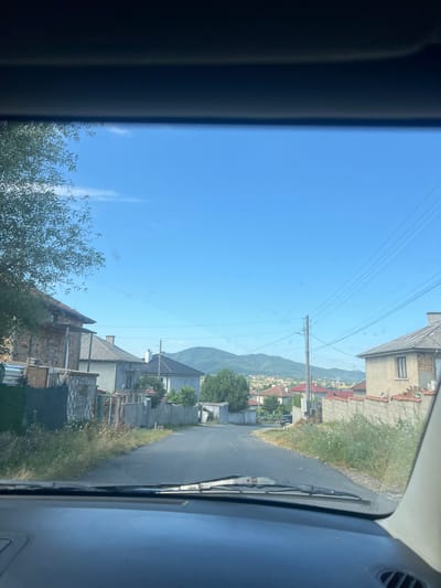 The image depicts a view from inside a vehicle, looking out through the windshield at a street scene in the city of Benkovski, Bulgaria. The road ahead curves slightly to the left and is bordered by houses and vegetation. On the left side of the road, there are several houses, some appearing to be under construction or in disrepair, with exposed brickwork and scaffolding. A large tree with sparse foliage is also visible on the left. On the right side, there is a fence made of concrete blocks topped with red tiles, and more houses further down the street. In the background, a town or village is spread out with red-tiled roofs, leading up to green, rolling hills under a clear blue sky. Power lines are strung across the sky, supported by a utility pole. The foreground shows the dashboard of the vehicle, with the windshield wipers visible. The lighting suggests it is daytime, likely late morning or early afternoon, with bright sunlight. There are no people clearly visible in the image.