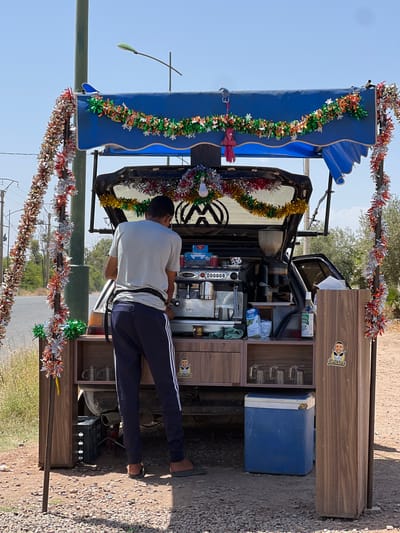 A man operates a mobile coffee stand constructed from the rear of a Volkswagen vehicle, Saâda, Morocco. The vehicle's back end is fitted with a wooden cabinet containing coffee-making equipment, including an espresso machine, cups, and various beverages. A blue cooler is positioned beside the cabinet. The vehicle's roof is partially covered by a blue awning. The stand is decorated with colorful metallic garlands. The man, wearing a white t-shirt and blue track pants, stands behind the cabinet operating the espresso machine. The scene is set outdoors on a dirt road. The weather appears sunny and clear. The mobile coffee stand is located on the side of a road in a rural setting with trees and vegetation visible in the background.