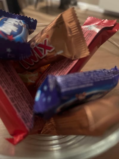 The image shows a close-up view of a variety of candy wrappers, some partially open, arranged in a clear glass bowl. The wrappers are of different colors including blue, red, gold, and pink. Some visible text on the wrappers includes "TWIX" and what appears to be Russian lettering. The background is blurred but suggests an indoor setting with a wooden floor and furniture. There are no people visible in the image. The location is Yastrebovskii, Russia.