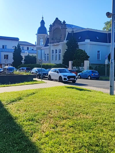 The image shows a street scene in Nový Jičín, Czechia, on a clear, sunny day. In the foreground, a patch of mowed grass is visible, casting a shadow from an unseen object to the left. Beyond the grass is a paved walkway and a street lined with parked cars. Several buildings are visible in the background, including a light-colored structure with a distinctive dark blue domed roof and ornate architectural details, possibly a public building or historical residence. Another building to the left is lighter in color with blue accents. Several cars are parked along the street, including a white SUV, a dark-colored hatchback, and a blue hatchback. A street sign with a "P" symbol, indicating parking, is visible on the right side of the image. The environment appears to be an urban or suburban setting, with clear blue sky overhead.