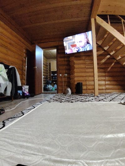 The image depicts the interior of a wooden log cabin, likely in Gorshki, Russia, given the context. The room is sparsely furnished, with a visible mattress in the foreground, suggesting it might be a bedroom or a multipurpose living space. A patterned rug covers part of the floor.
The walls and ceiling are constructed from horizontally stacked logs, characteristic of traditional Russian rural architecture. A television is mounted on one of the log walls, displaying a young person with short blonde hair wearing a plaid shirt. Adjacent to the television is a wooden staircase leading to an upper level or loft.
To the left of the television, several items of clothing hang on a rack, with bags and other items piled on the floor beneath them. A doorway is visible in the background, leading to another room or area, which appears to contain shelves with various objects and some larger bags. A small white fan and a black cylindrical object, possibly a speaker or a heater, are placed on the floor near the wall.
Electrical wires are visible on the floor and wall, indicating the presence of lighting and power outlets. The overall lighting in the scene is dim, with light sources originating from the television and the open doorway. The environment appears functional rather than ornate, suggesting a lived-in, possibly rustic dwelling.