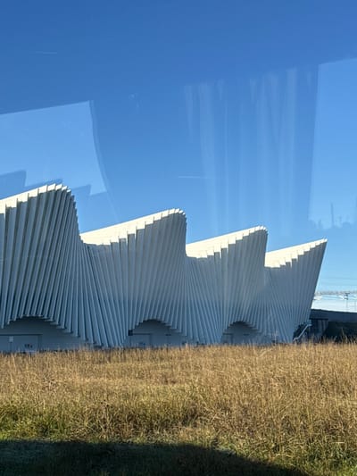 The image displays a portion of the Reggio Emilia AV Mediopadana high-speed railway station in Reggio nell'Emilia, Italy. The prominent subject is the station's distinct architectural facade, characterized by multiple undulating, wave-like structures composed of white, vertically oriented, twisting fin-like elements. These structures exhibit a repetitive pattern of peaks and valleys, extending horizontally across the frame. At the base of the white facade, several recessed, dark rectangular openings are visible, consistent with entryways or service access points. The foreground consists of a dense field of dry, golden-brown and light-yellow grass, occupying approximately the lower third of the image. The upper two-thirds of the image depict a clear, bright blue sky, with visible reflections and minor distortions suggesting the photograph was taken through a window, possibly from a vehicle. In the far right background, a faint structural element, consistent with a bridge or industrial infrastructure, is discernible. No individuals or active events are present in the scene.