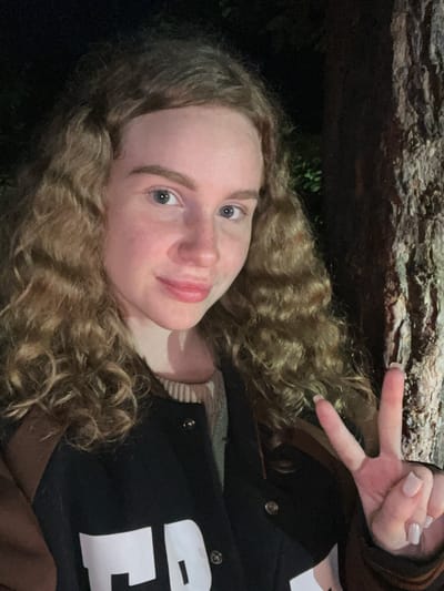 A person with curly blonde hair and blue eyes is posing for a selfie. They are making a peace sign with their right hand. They are wearing a black jacket with white lettering, possibly a letterman jacket. The background is dark, but shows a tree trunk, possibly indicating they are outdoors in a wooded area. There are no indications in this image that definitively establish that this image was taken in Krasnyi Jar, Russia.
